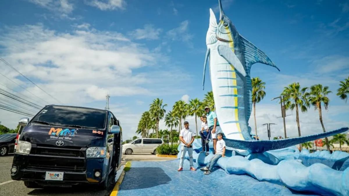 A branded MyM Travel & Tours van parked beside the blue marlin sculpture, with palm trees behind